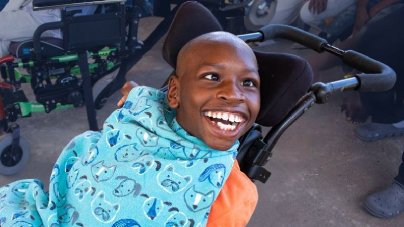 Mark is a young boy in a wheelchair wearing an orange shirt and blue apron. He is smiling up at a friend.